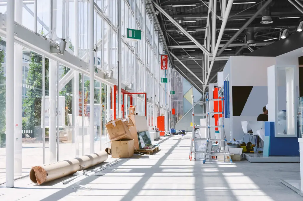 A Busy Exhibition Build Scene With Boxes And Materials On The Floor Adjacent To Two Ladders.