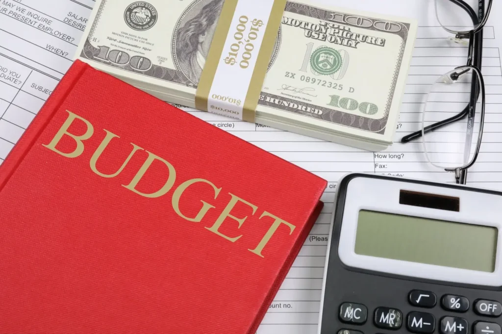 A red book titled "budget" placed next to a calculator, a wad of dollar bill, and a pair of glasses.