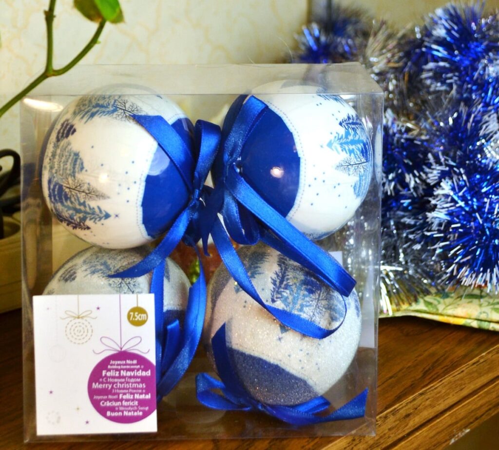 Four blue and white Christmas baubles packaged in a clear plastic box with tinsel in the background. 