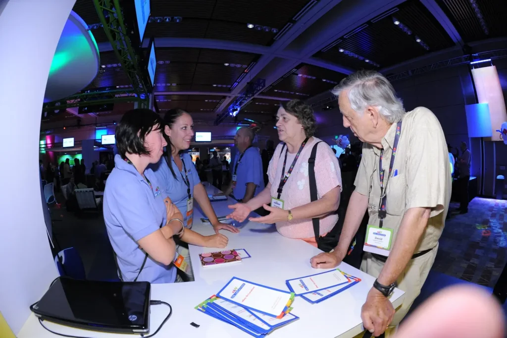 Booth staff interact with two guests at a table during an exhibition. 