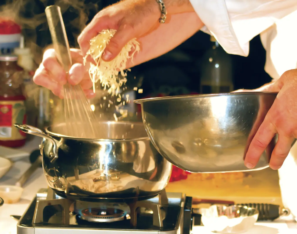 One chef drops grated cheese into a pot on the stove while the other chef whisks. 